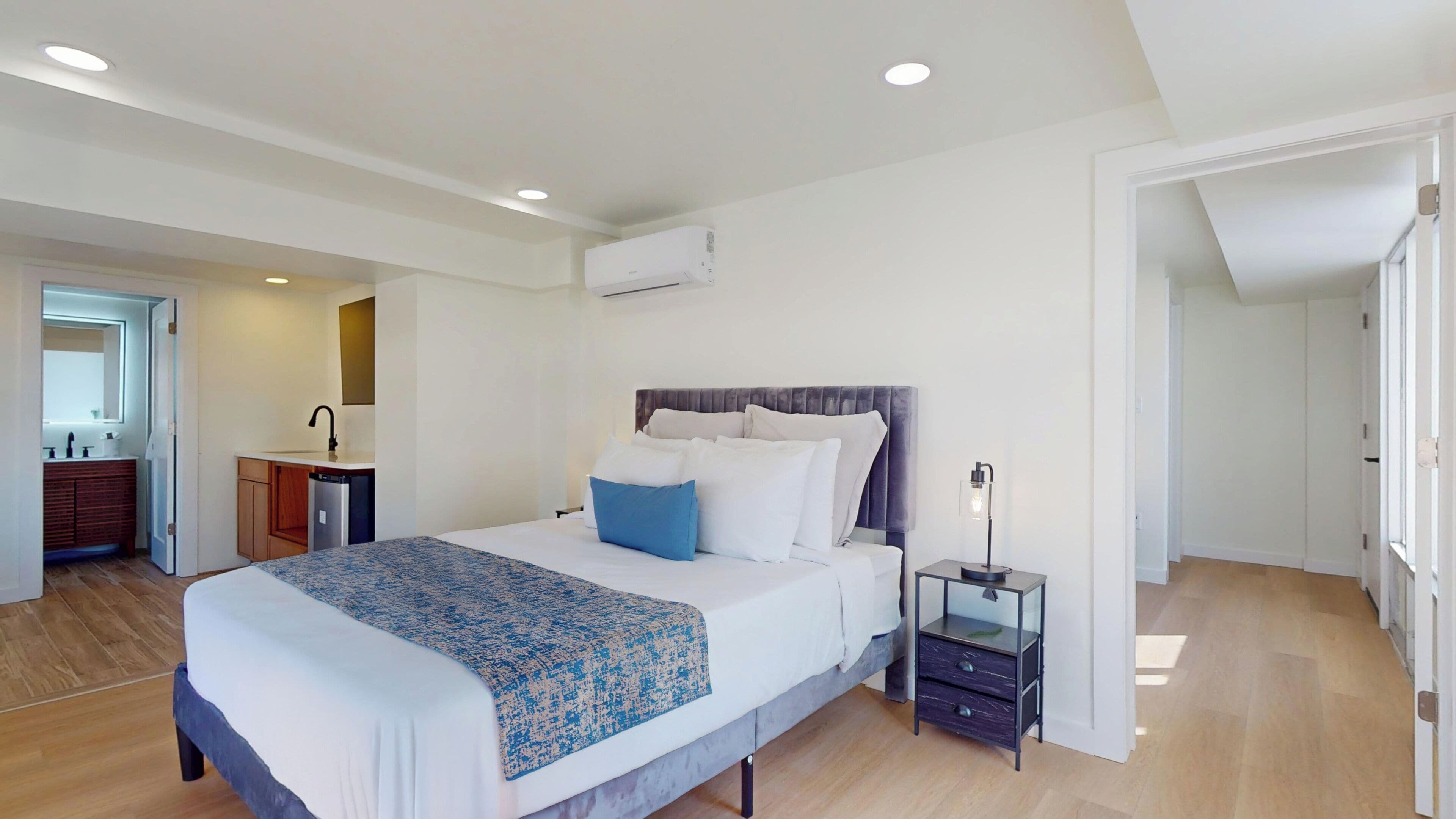 Una habitación de hotel moderna con una cama grande, una mesita de noche, un escritorio integrado con una silla, un espejo y un equipo de aire acondicionado.