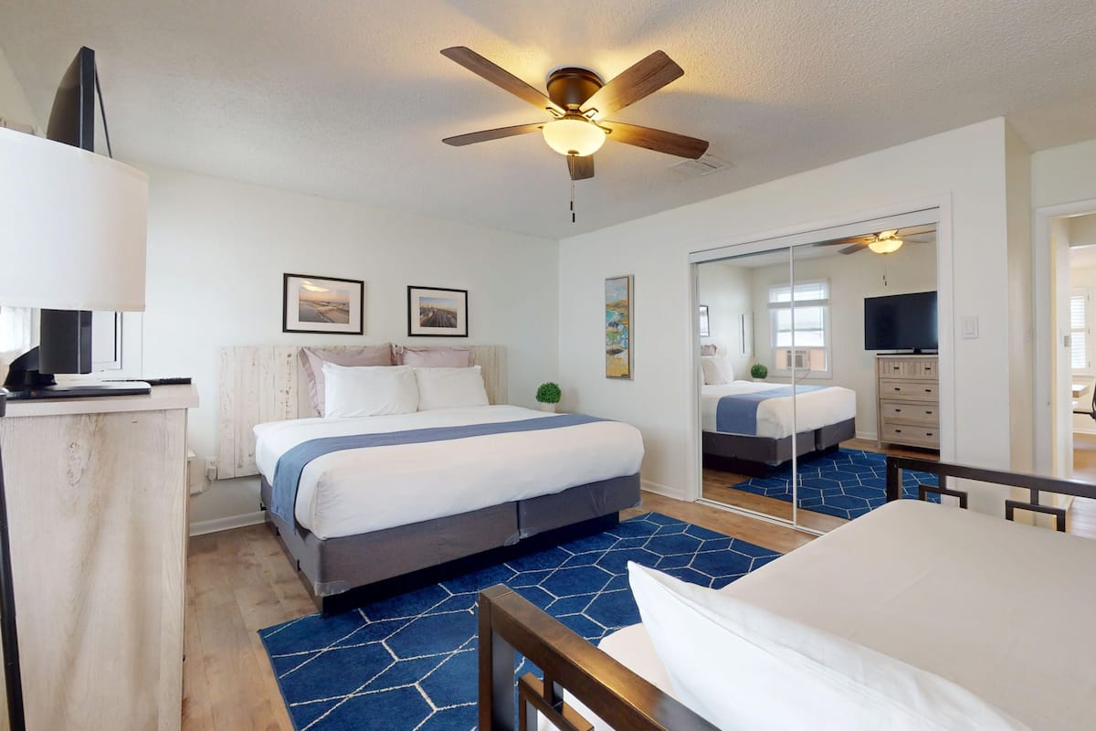 A hotel room with a bed, a desk, a ceiling fan, a TV, and a large mirror.