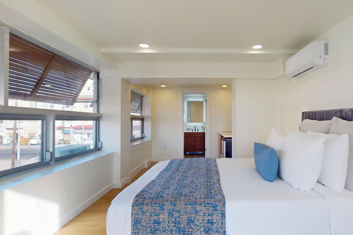 A hotel room with a large bed, a window with blinds, a bathroom with a mirror, and an air conditioning unit.