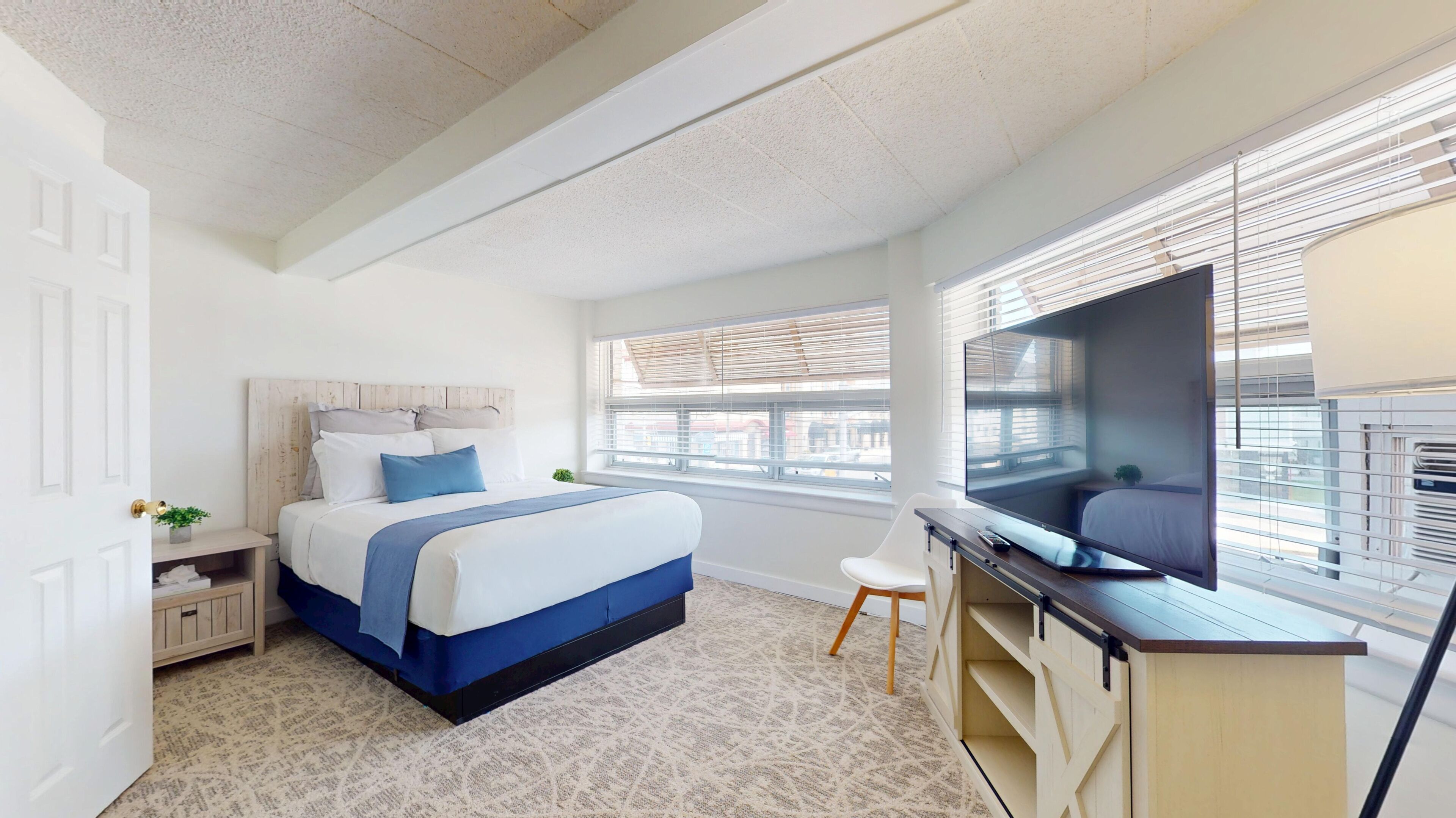 A bedroom with a bed, a TV, a desk, and a window with blinds.