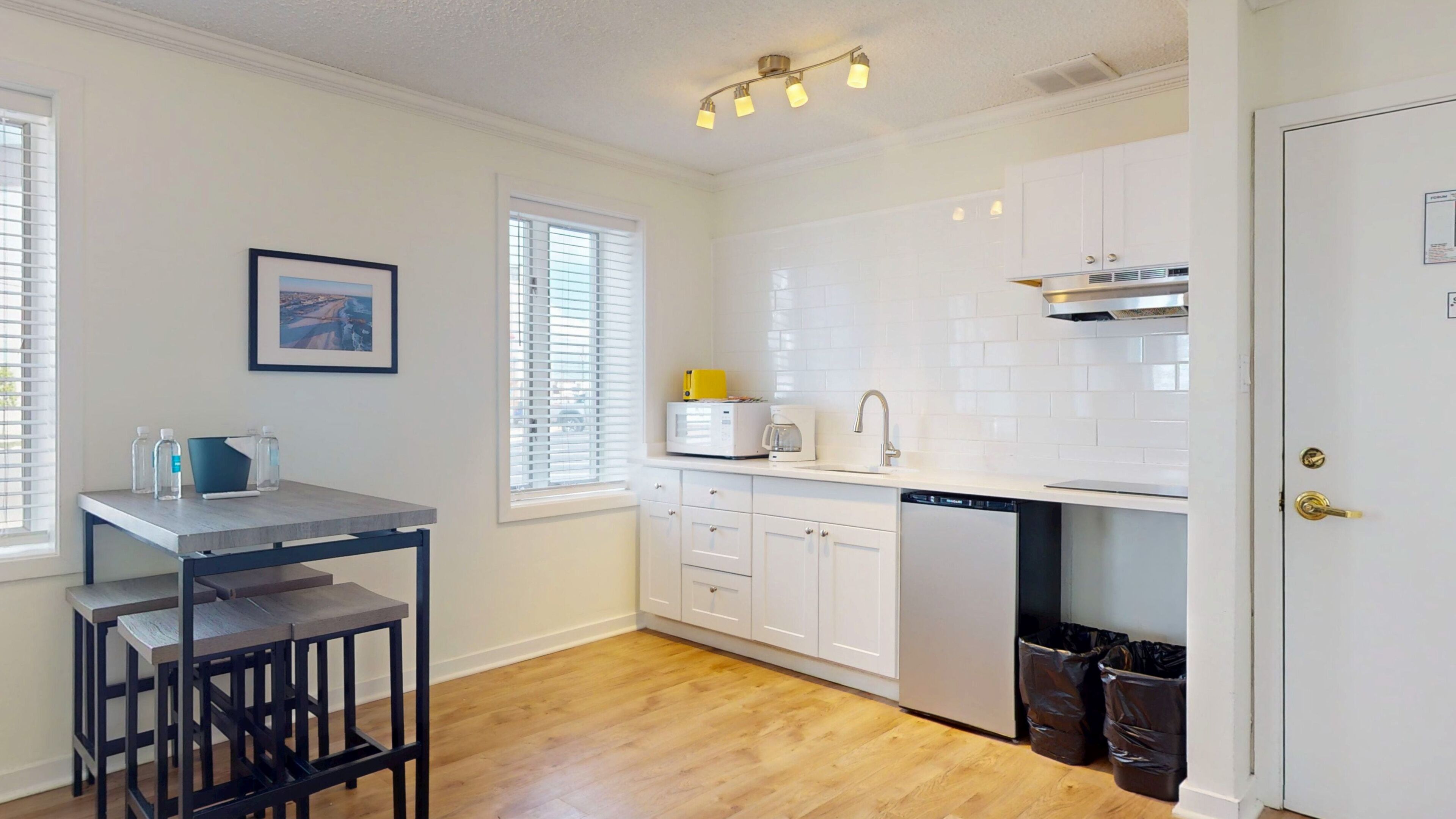 A modern kitchen with white cabinets, a microwave, and a coffee maker. There is a dining table with chairs and a window with blinds.