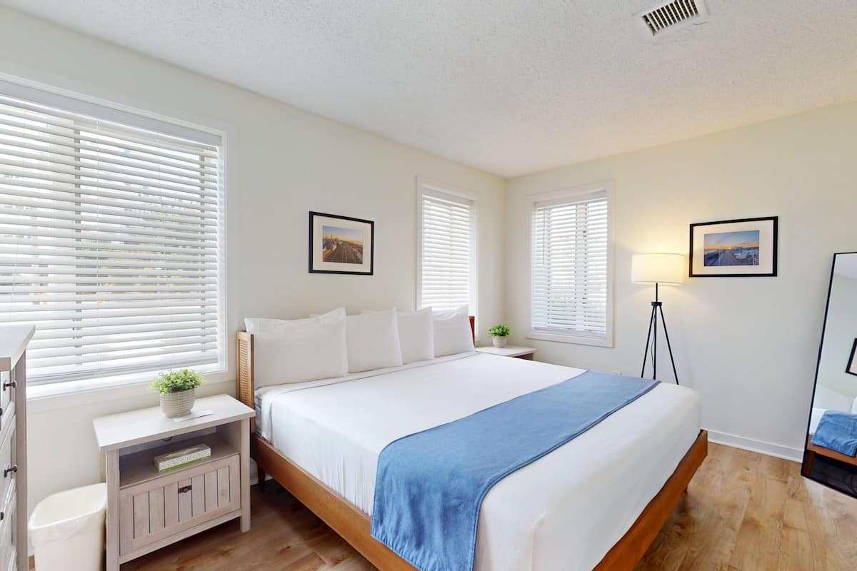 A bedroom with a bed, bedside tables, a lamp, a mirror, and windows with blinds.
