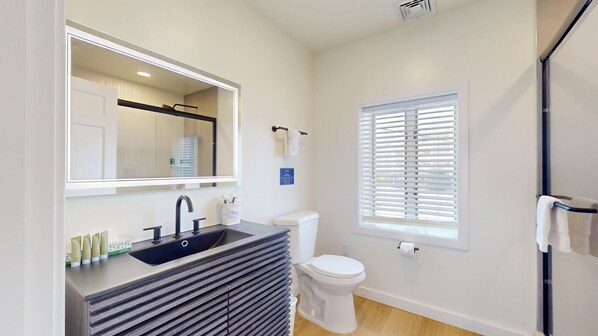 Un baño moderno con un espejo grande, un lavabo con artículos de aseo, un inodoro y una ventana con persianas.