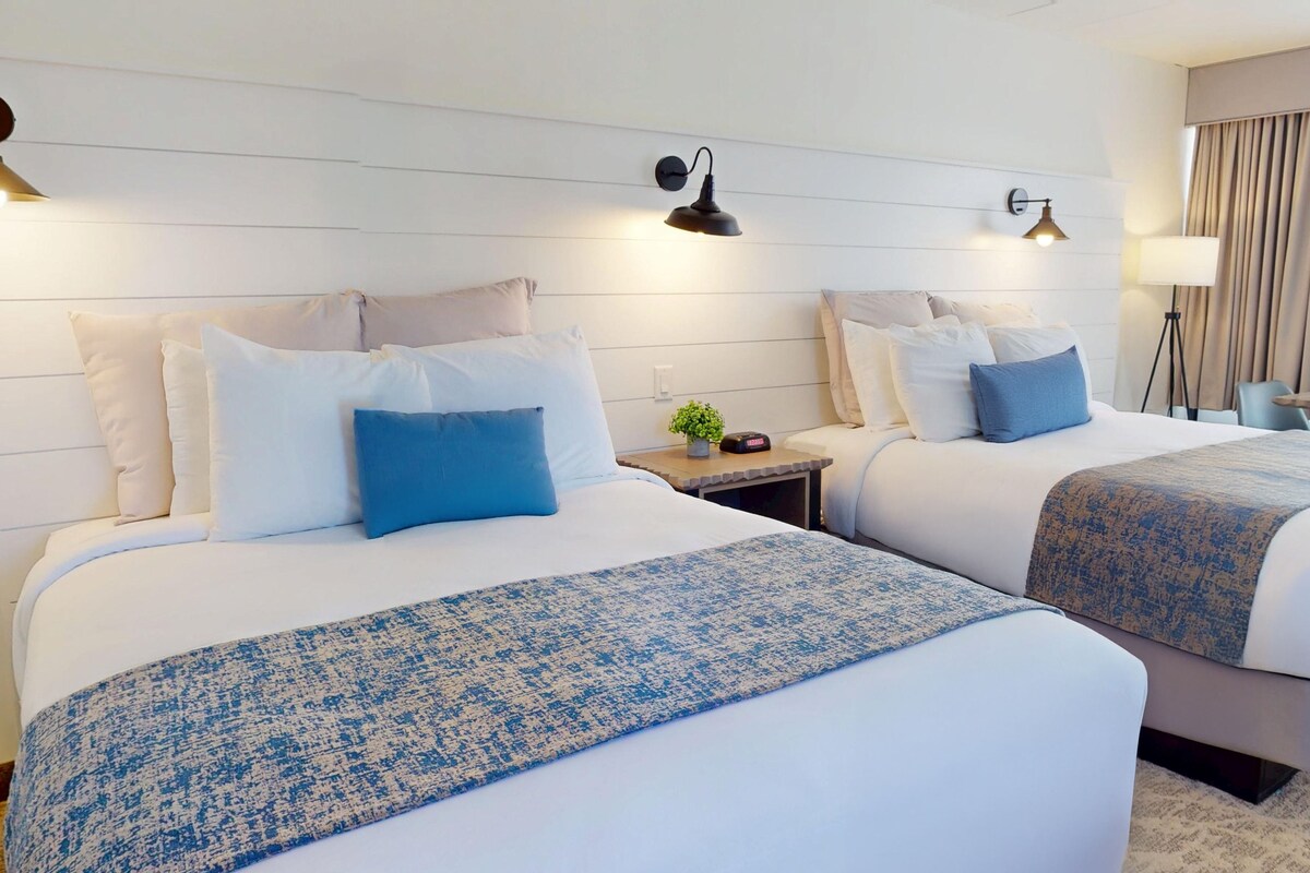 A hotel room with two beds, a bedside table with a clock, a lamp, and a view of the outside through a window.