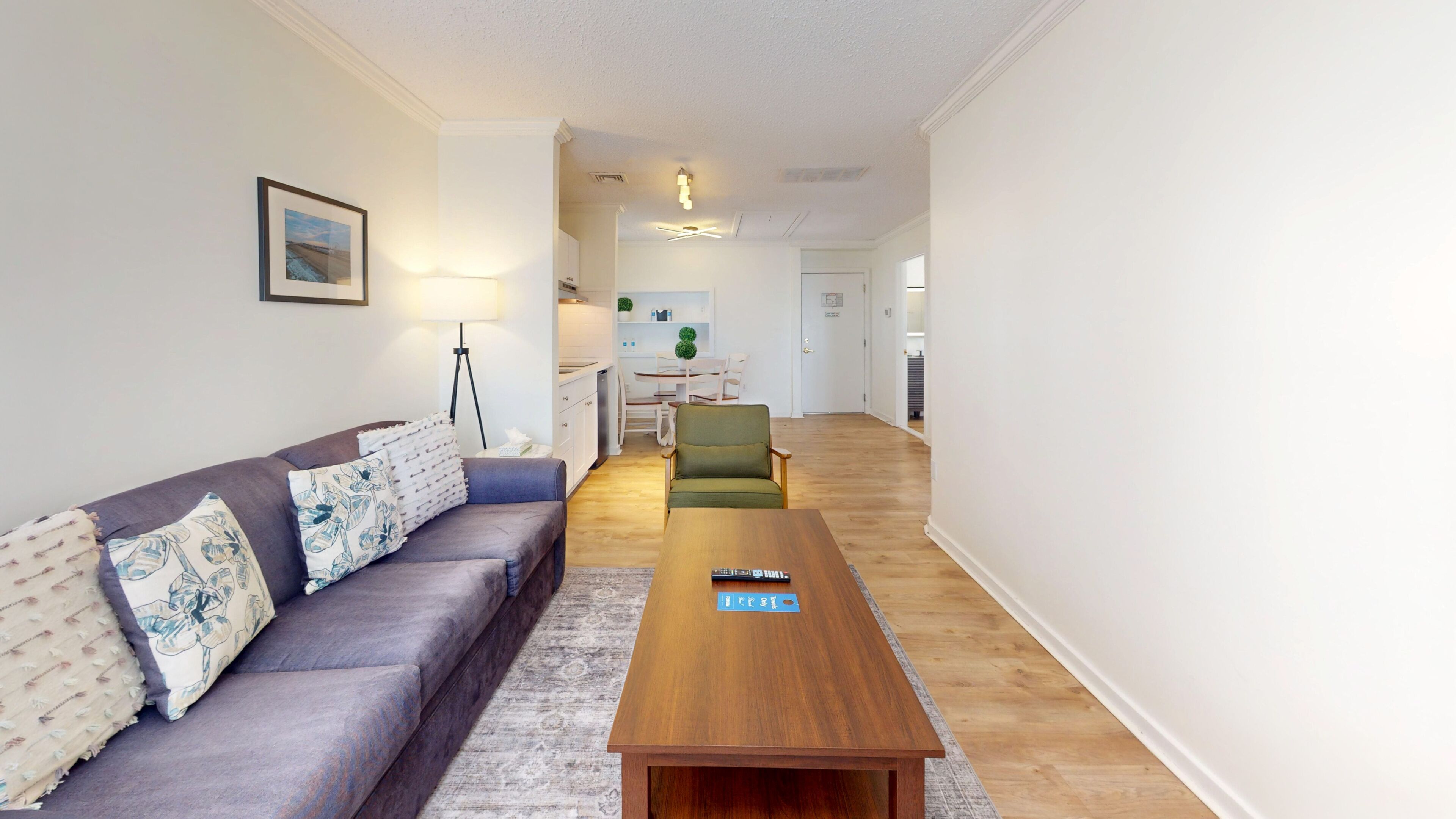 Una sala de estar con un sofá, una mesa de centro y una silla. Se observa una cocina al fondo.