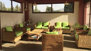 Un espacio de estar al aire libre cubierto con mobiliario de mimbre y cojines verdes, amplias ventanas que ofrecen vistas a árboles y colinas.