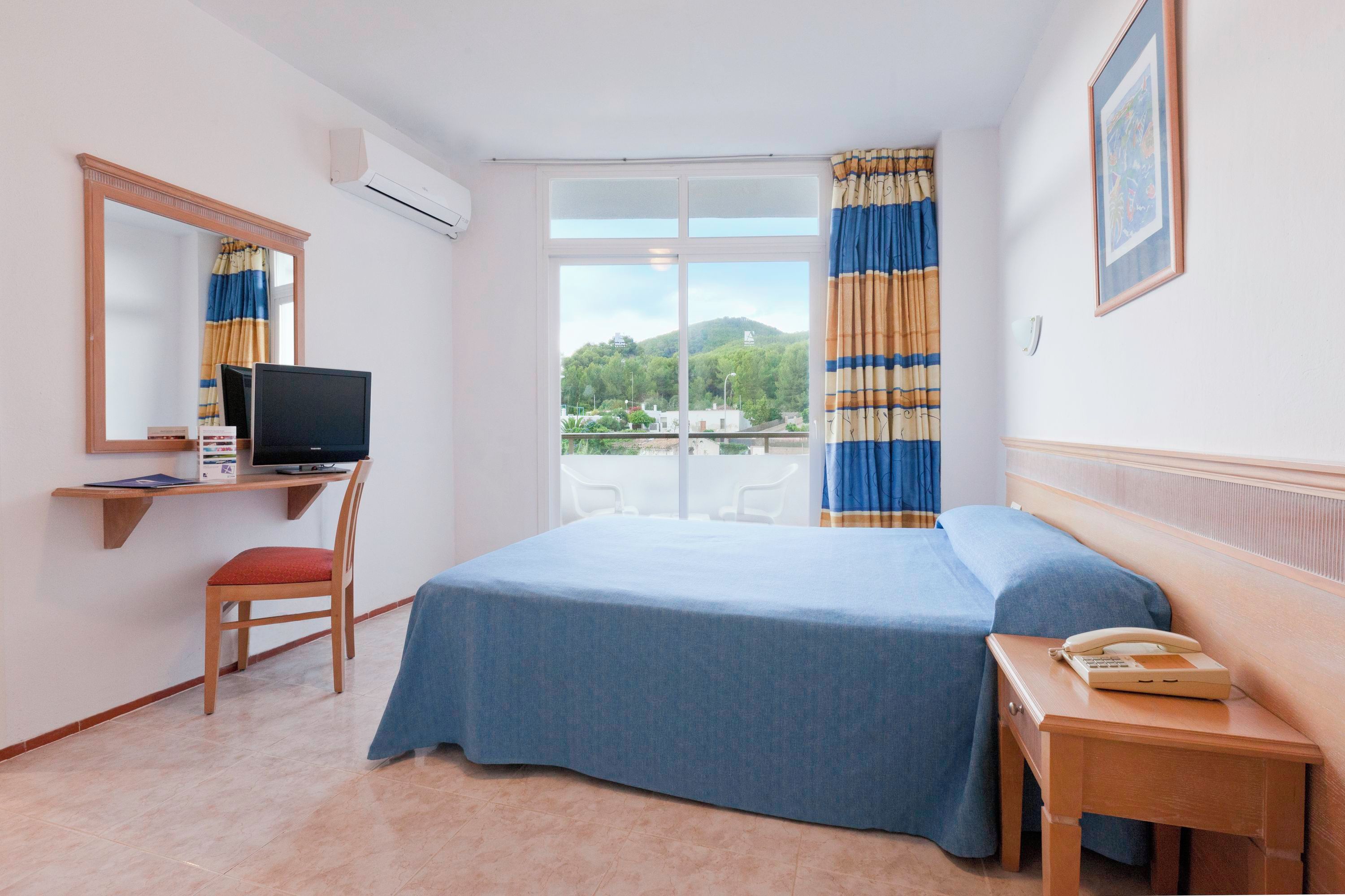 A hotel room with a bed, a desk with a computer, a chair, a telephone, and a window with curtains.