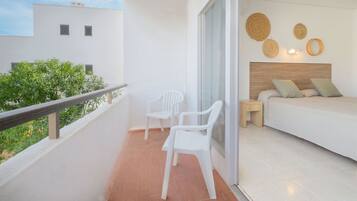 Een balkon met twee witte plastic stoelen, een houten nachtkastje, een bed met houten hoofdbord en een wand met wanddecoratie van geweven materiaal.