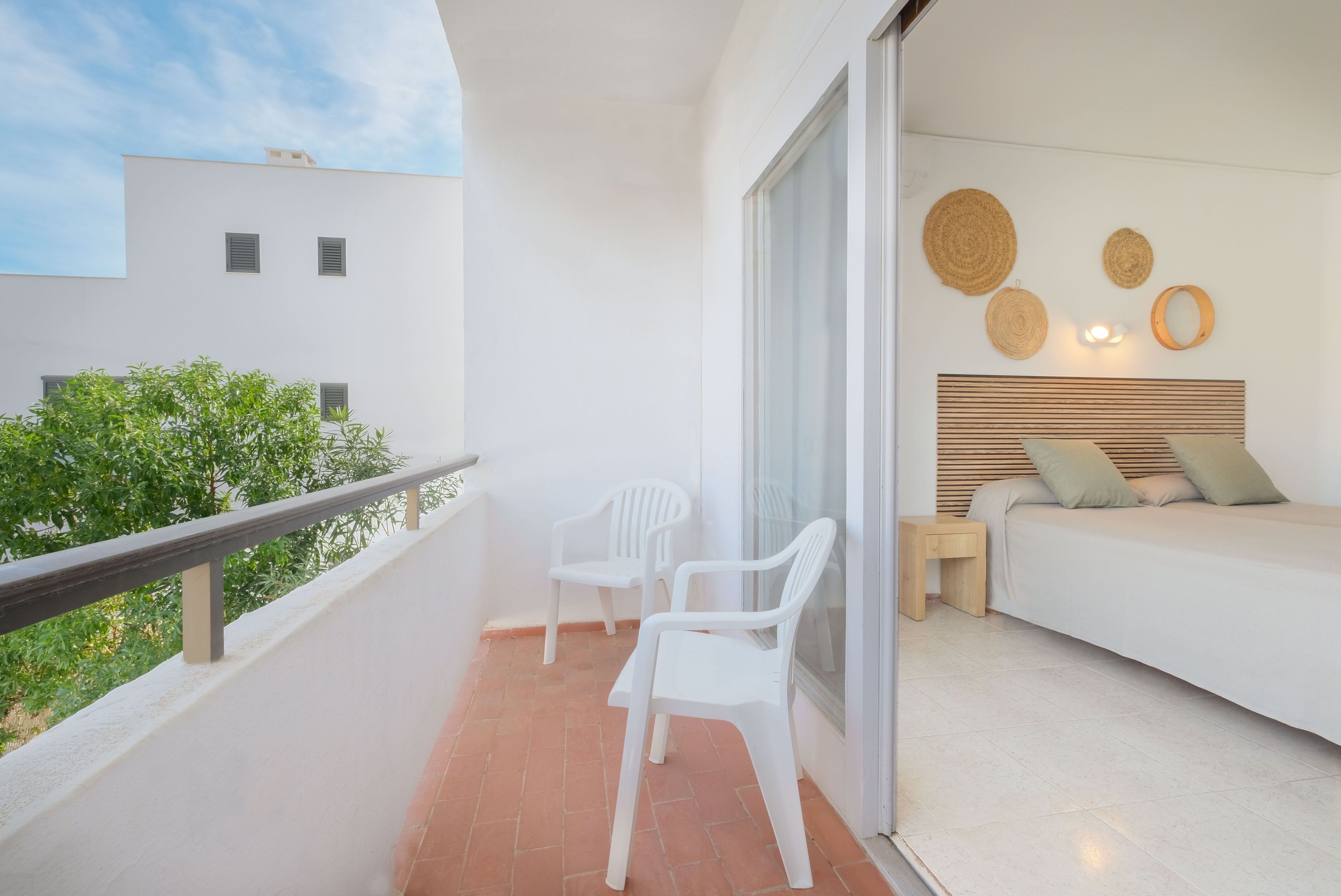 A balcony with two white plastic chairs, a wooden nightstand, a bed with a wooden headboard, and a wall with woven wall art.
