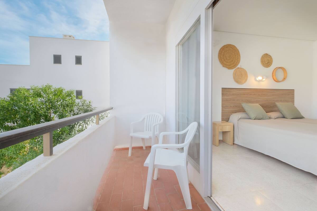 Un balcon avec deux chaises en plastique blanc, une table de chevet en bois, un lit avec une tête de lit en bois et un mur orné d’une décoration murale tissée.