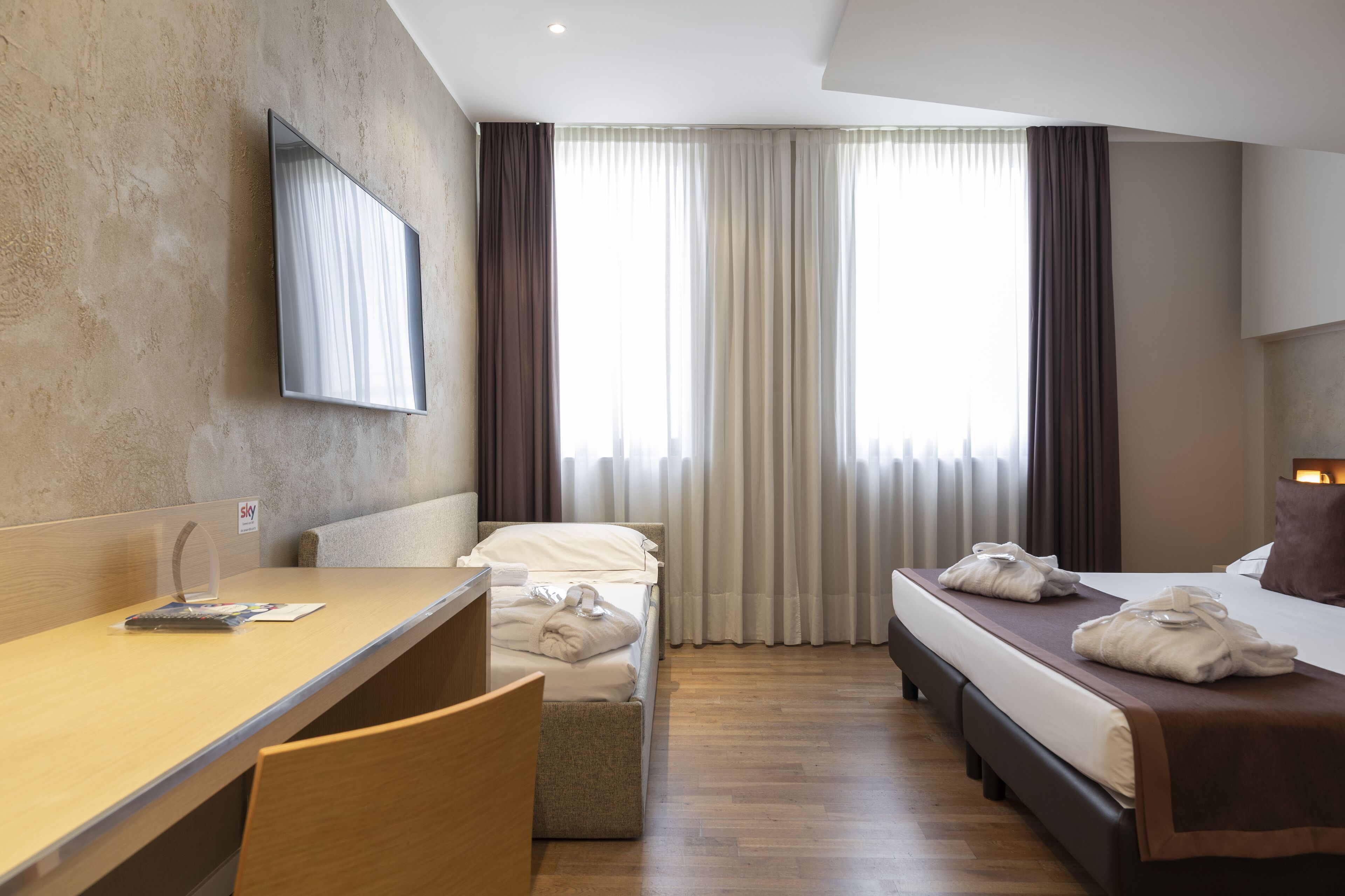 A hotel room with a bed, a desk, and a chair. There is a window with curtains, a flat-screen TV mounted on the wall, and a sign that says "sky".
