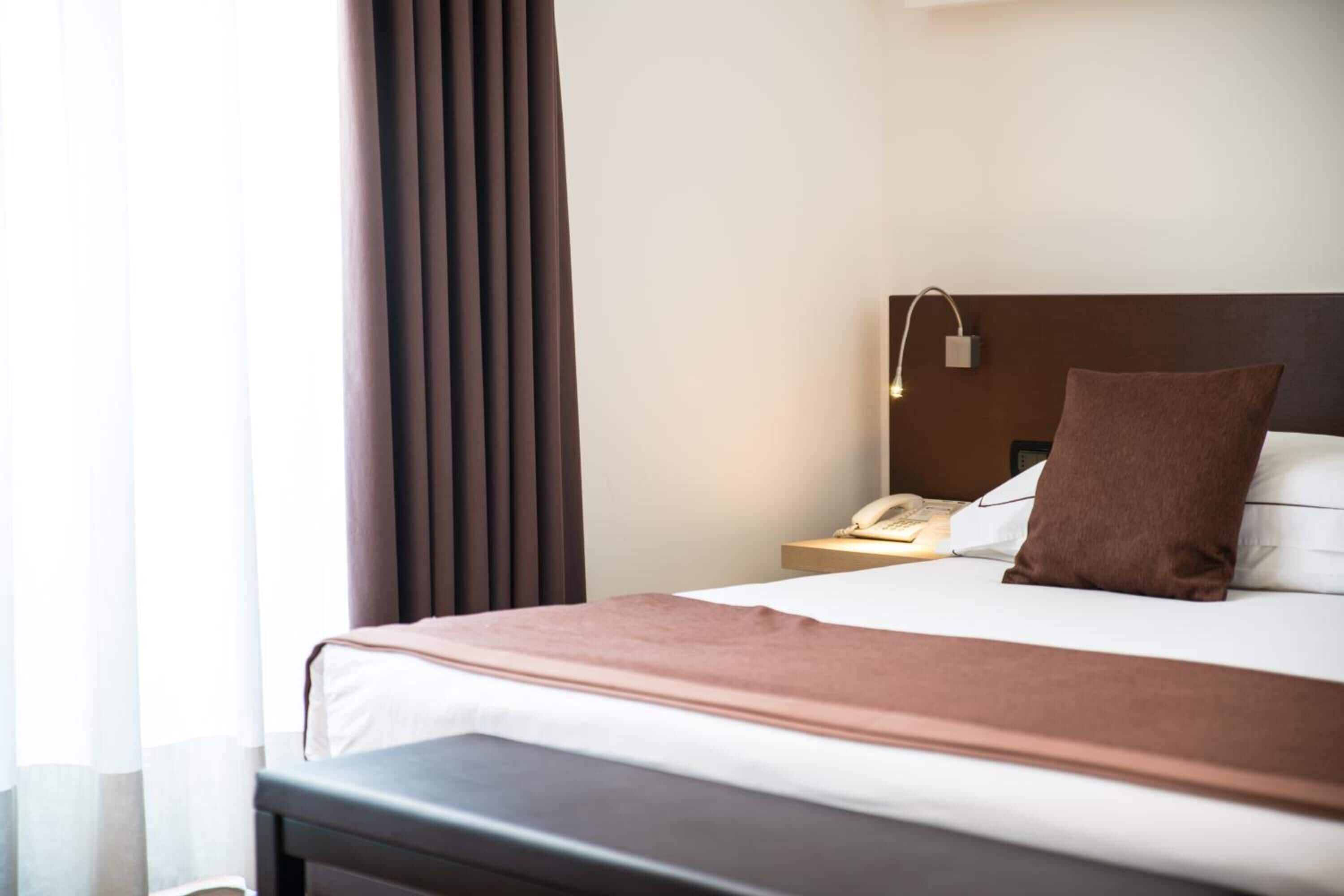 A hotel room with a bed, a bedside table with a lamp, a telephone, and a brown pillow.