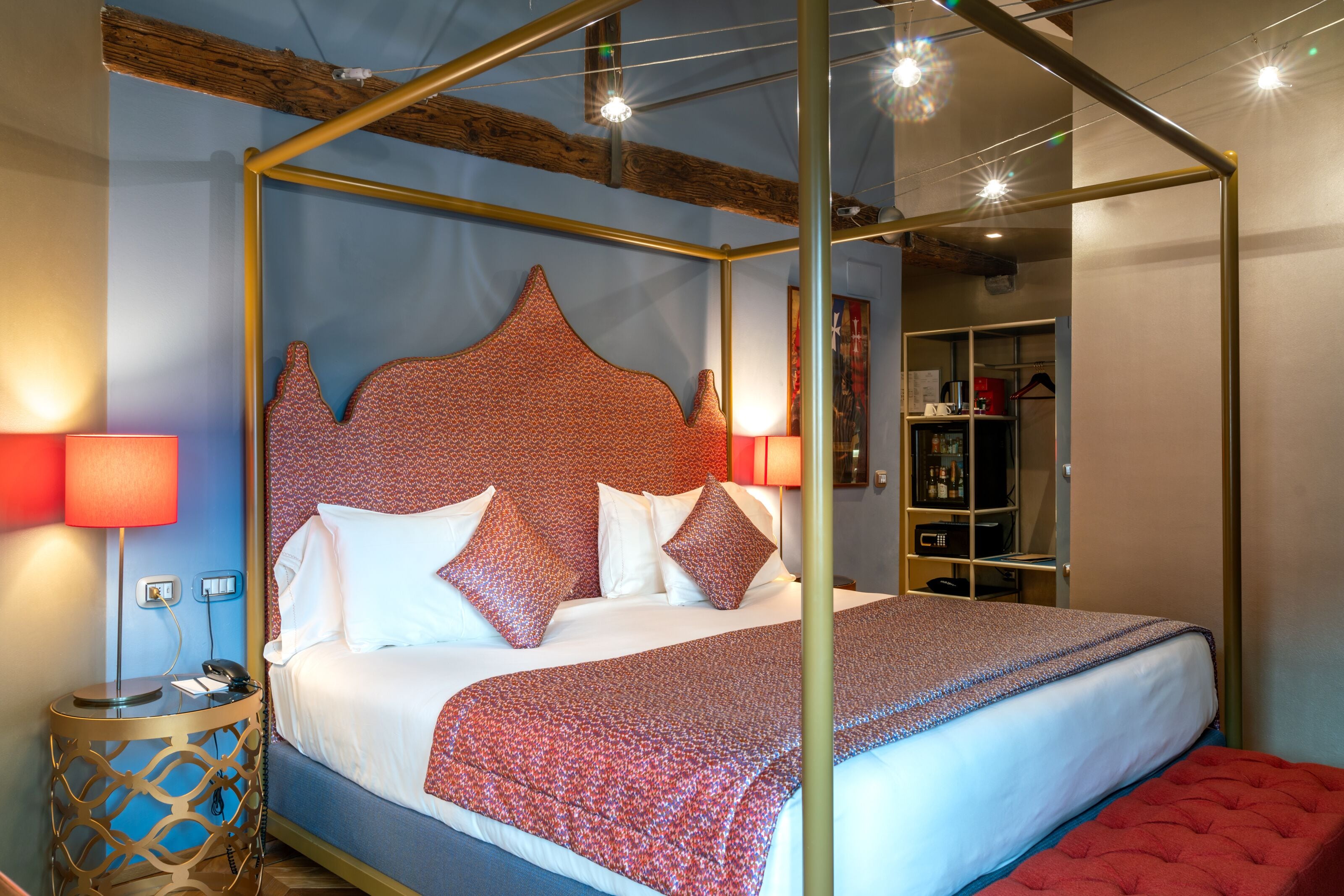Ein Bett mit Baldachin, ein gemustertes Kopfteil, Nachttischlampen und eine rote Polsterbank.