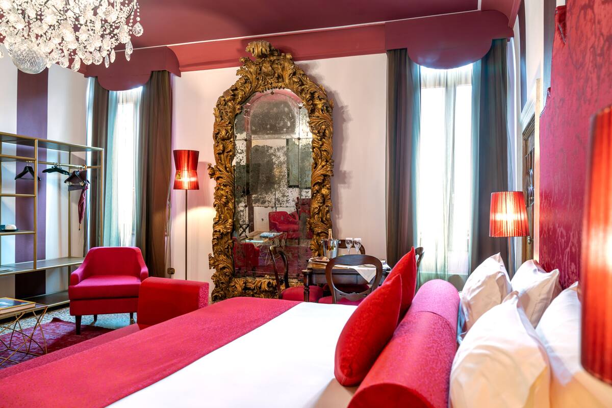 A hotel room with a large bed, red armchairs, a mirror with an ornate frame, and a chandelier.