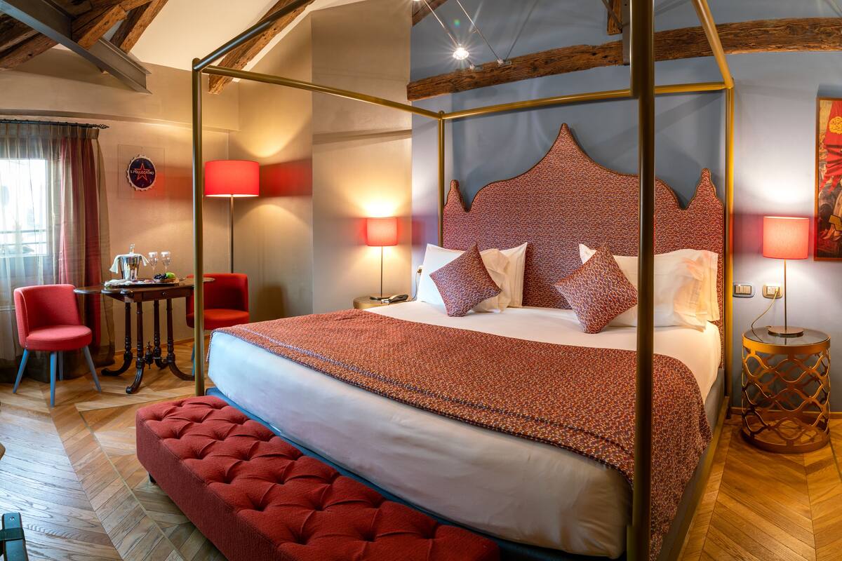 A four-poster bed with a red and white patterned bedspread, a red upholstered bench, and a wooden floor with herringbone pattern.