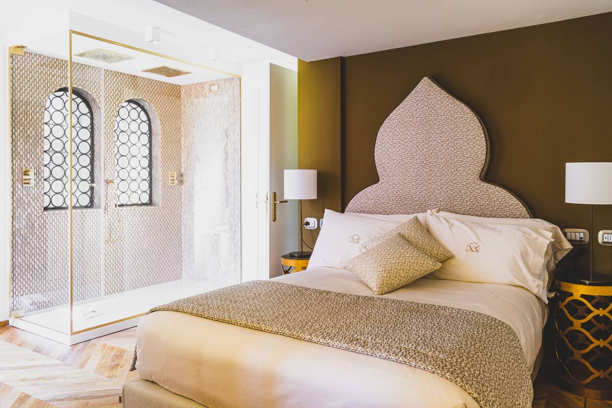 A bedroom with a large bed, two bedside tables, a nightstand, a decorative wall, and a glass door leading to a bathroom.