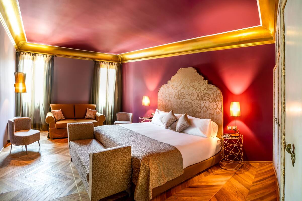 A spacious hotel room with a large bed, a sitting area with a sofa and chairs, and a wooden herringbone floor.