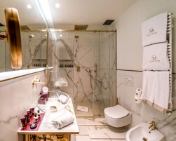 Un baño moderno con ducha de vidrio, inodoro blanco, lavamanos con grifo dorado y tocador con artículos de aseo.