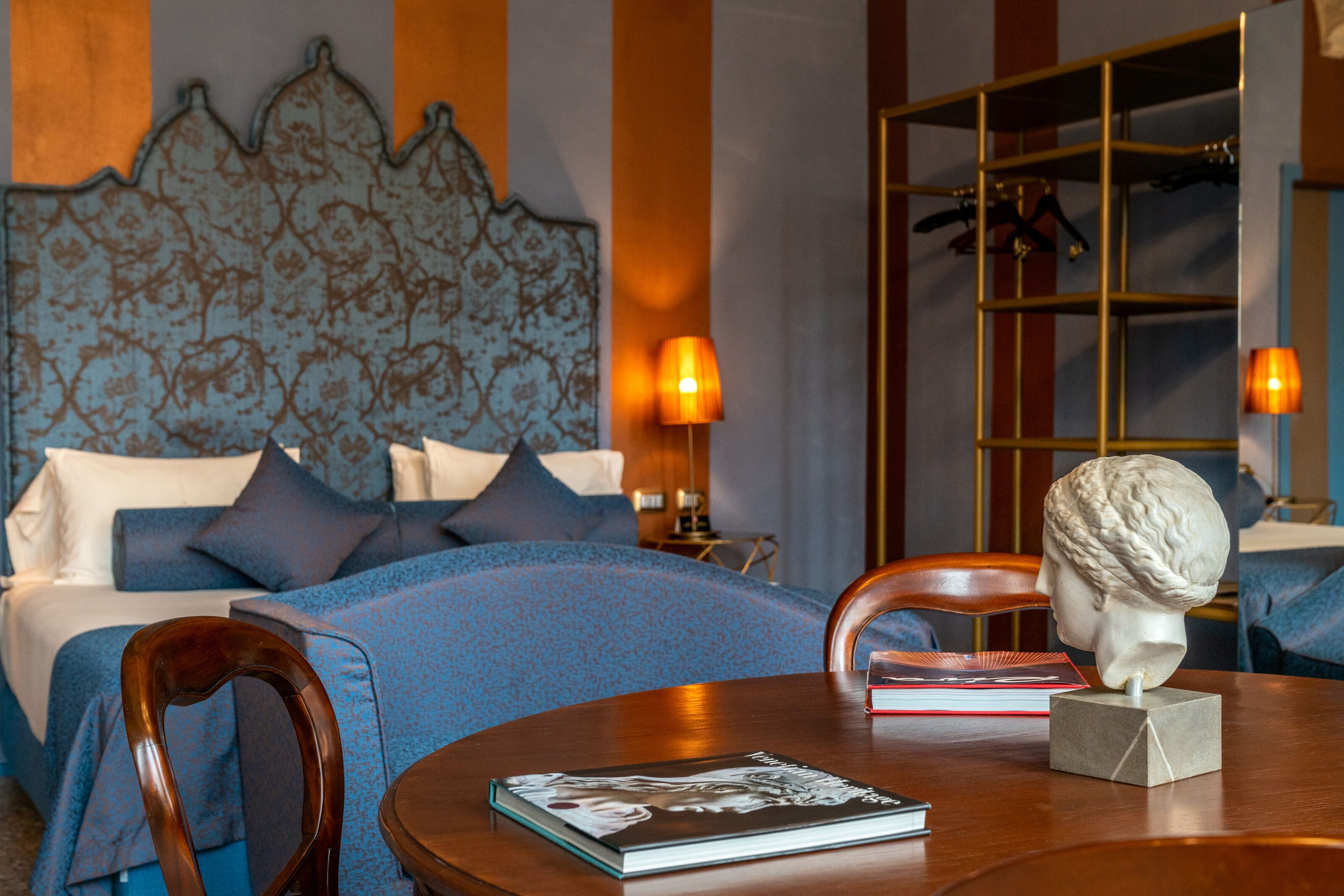 A hotel room with a bed, a wooden chair, a table with a book and a bust sculpture, and a lamp.