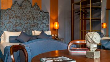 A hotel room with a bed, a wooden chair, a table with a book and a bust sculpture, and a lamp.