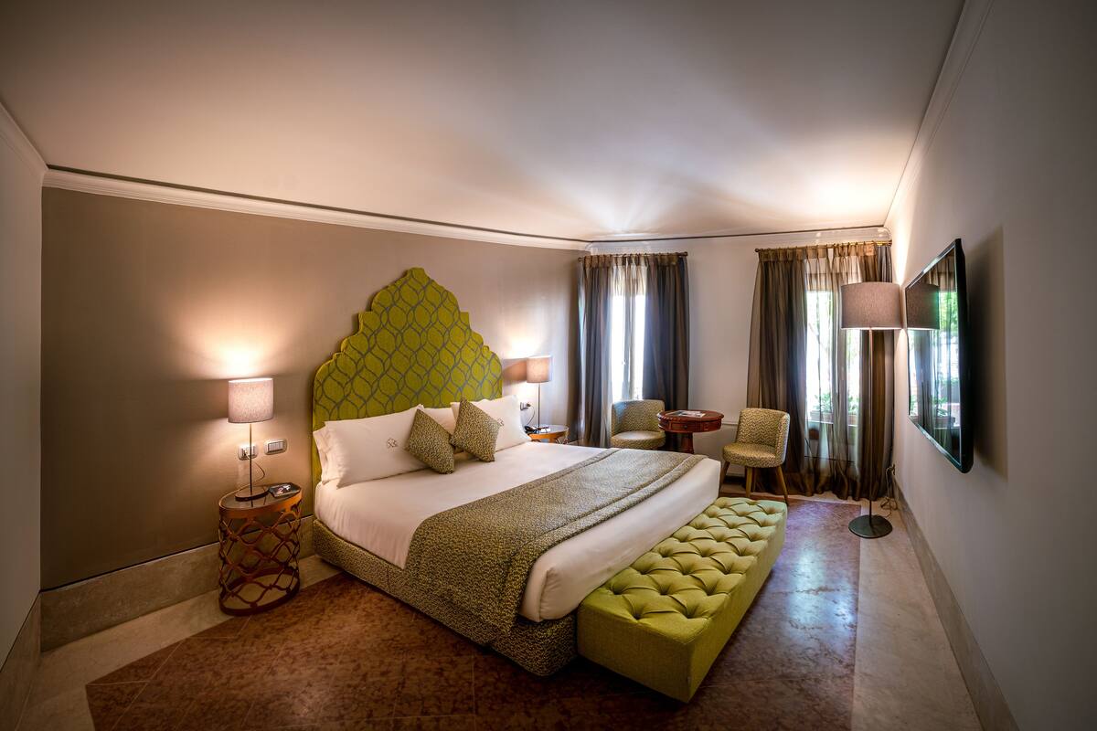 A hotel room with a large bed, two chairs, a round table, a TV, and a lamp.