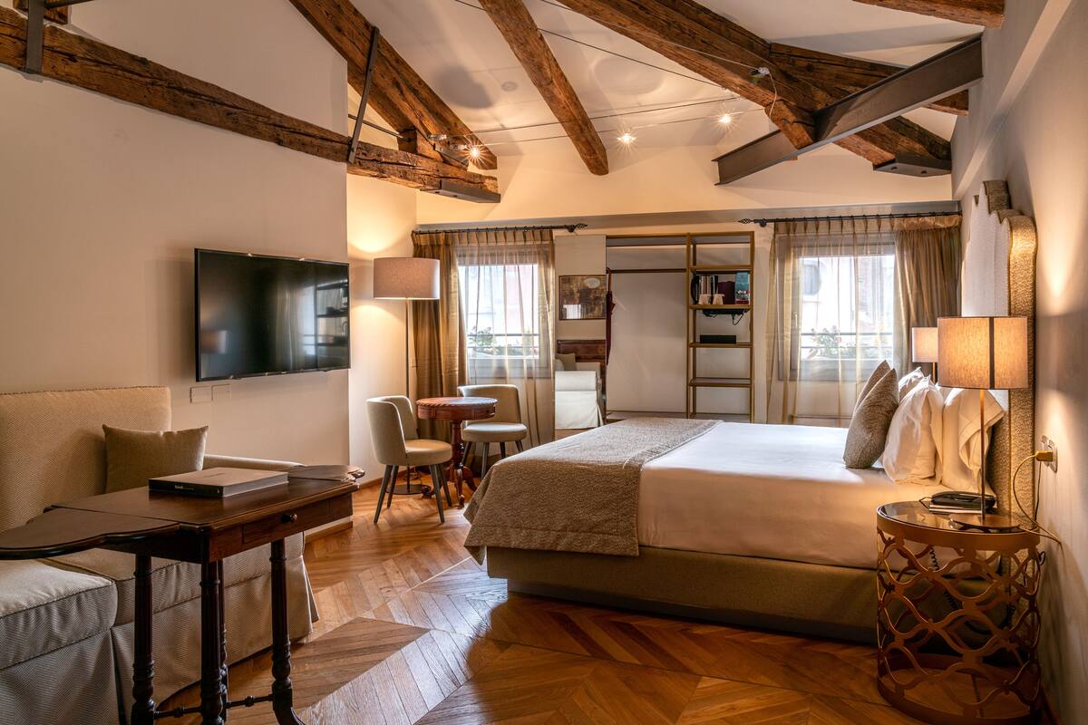 A well-lit bedroom with a bed, bedside table, lamp, and a TV mounted on the wall.