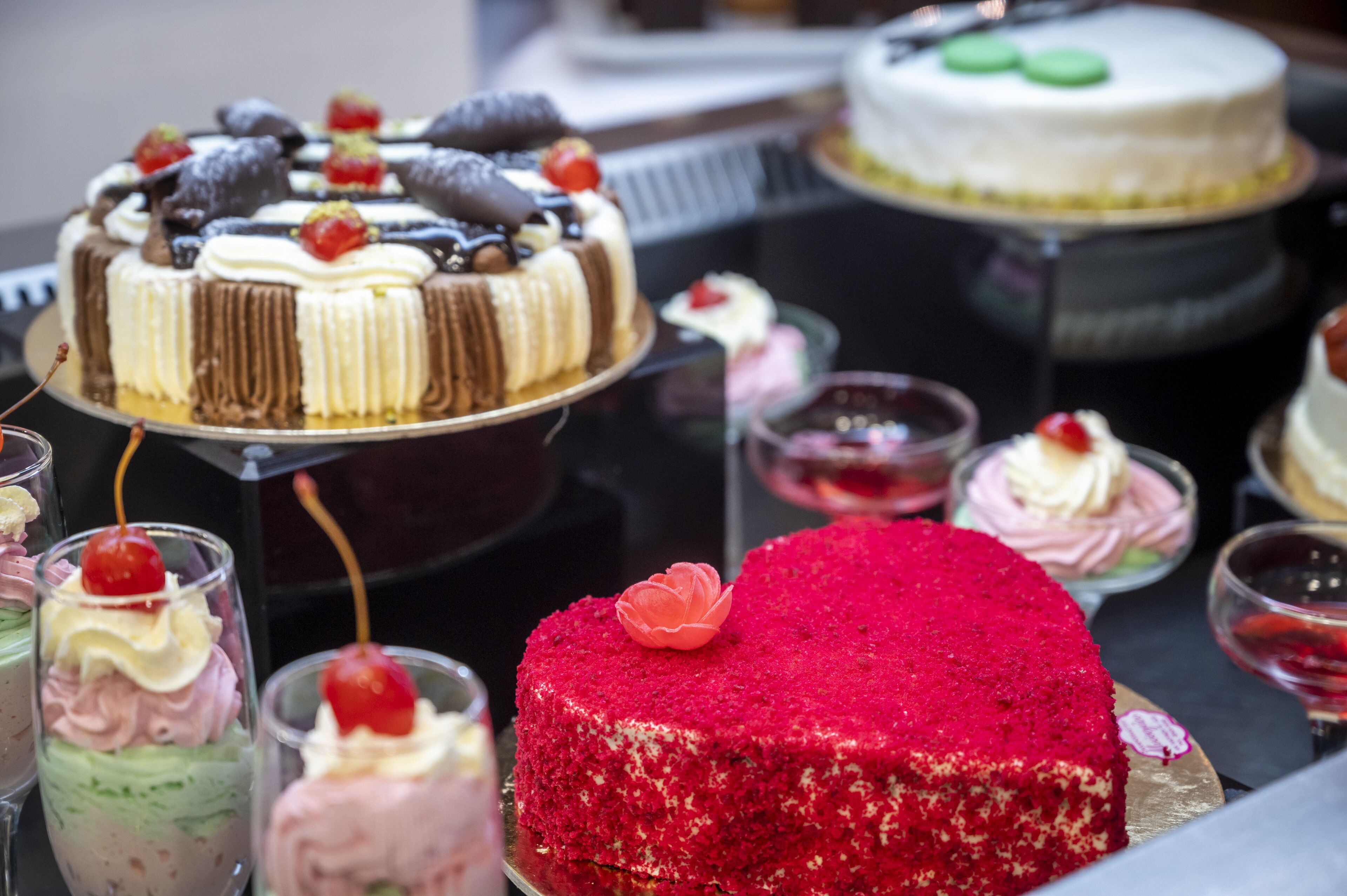 A variety of cakes and desserts on display, including a prominent red velvet cake with a cherry on top.