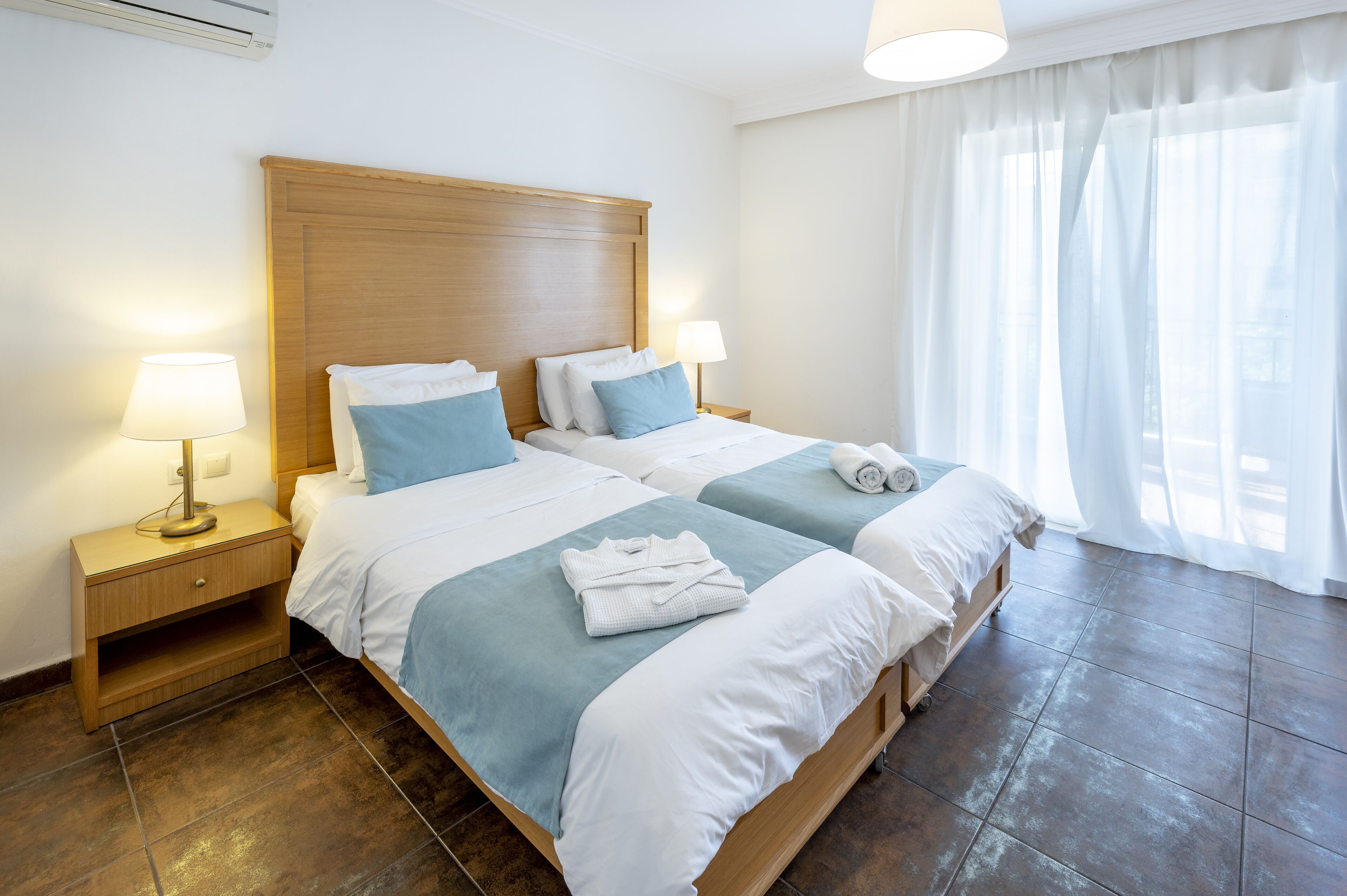 Una habitación de hotel con dos camas, cabecera de madera, lámparas de noche, una ventana con cortinas traslúcidas y suelo de baldosas.