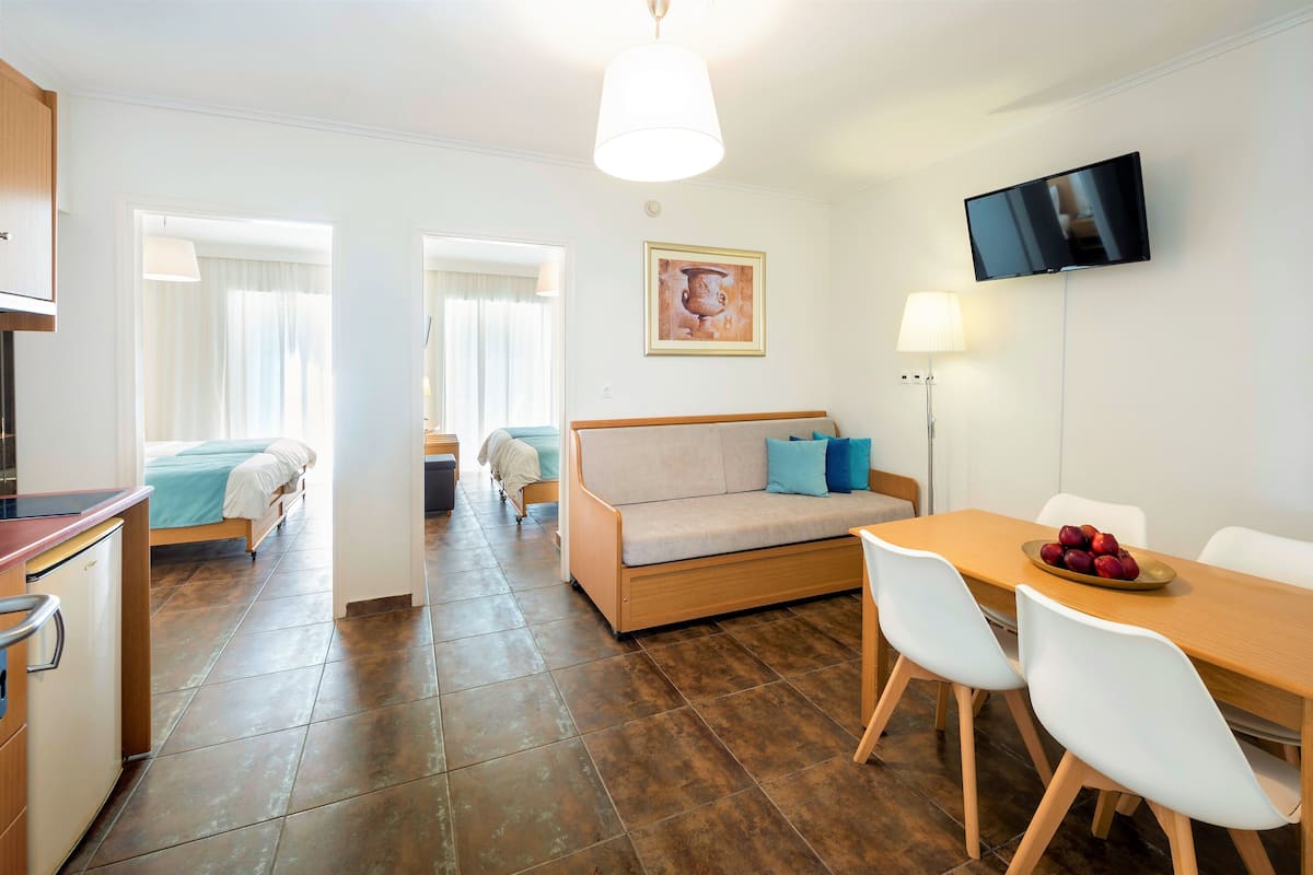 A modern hotel room with a kitchenette, a sofa, a dining table with chairs, a flat-screen TV, and a painting on the wall.