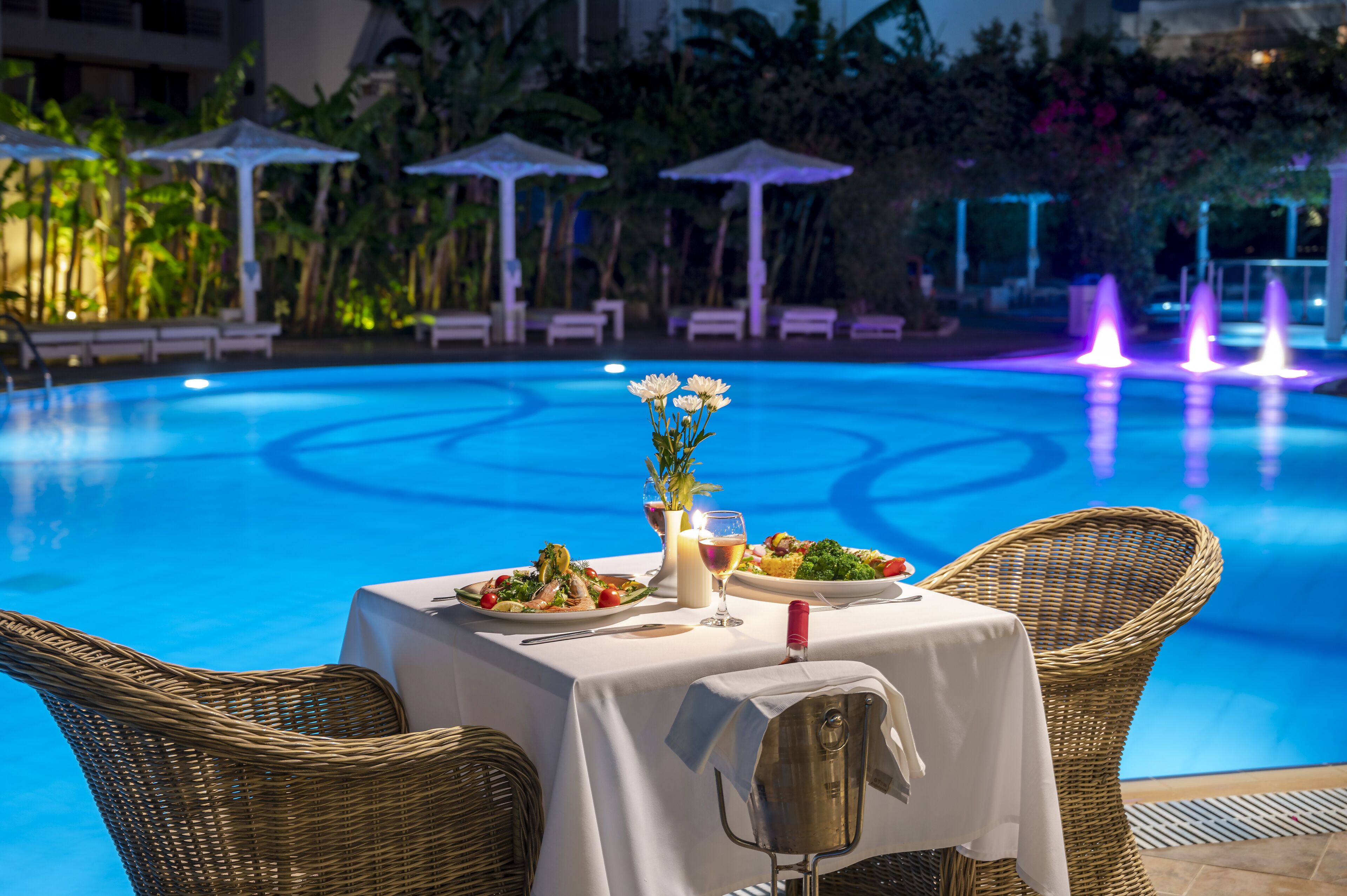 A dining table with a candle, a glass of wine, and a plate of food by a poolside.
