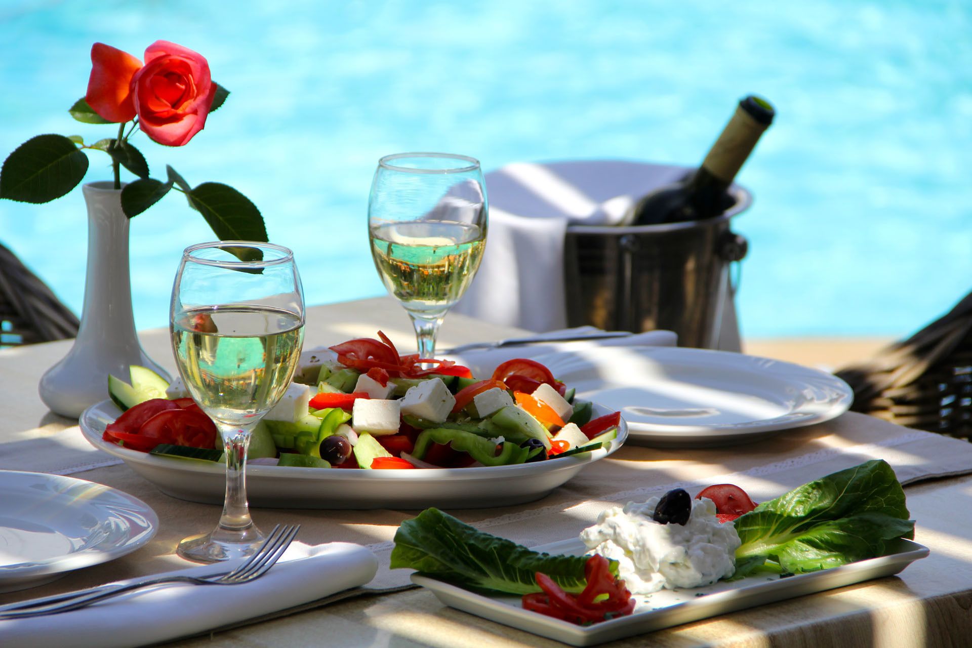 Una mesa servida con una ensalada, dos copas de vino blanco, una botella de champán y un florero con una rosa roja.