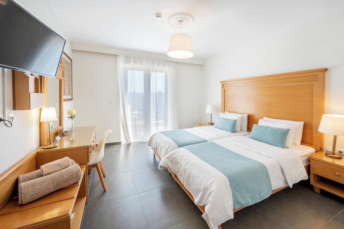 A hotel room with two beds, a desk with a chair, a TV, and a large window with curtains.