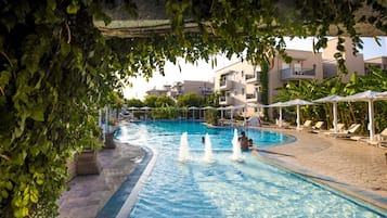 Ein Pool mit Wasserspielen, umgeben von Liegestühlen und Sonnenschirmen, mit Gebäuden im Hintergrund.
