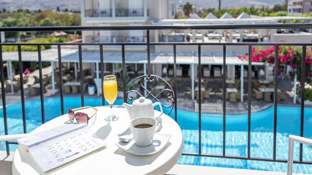 A table with a book, a cup of coffee, and a glass of orange juice on a balcony overlooking a swimming pool. - Peridis Family Resort (Kos)