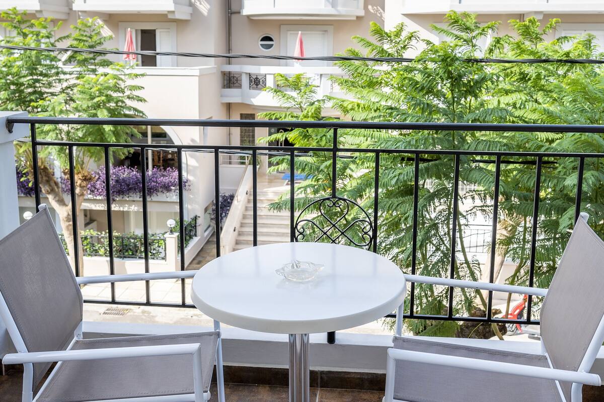 A balcony with a round table and two chairs, overlooking a residential area with white buildings and greenery.