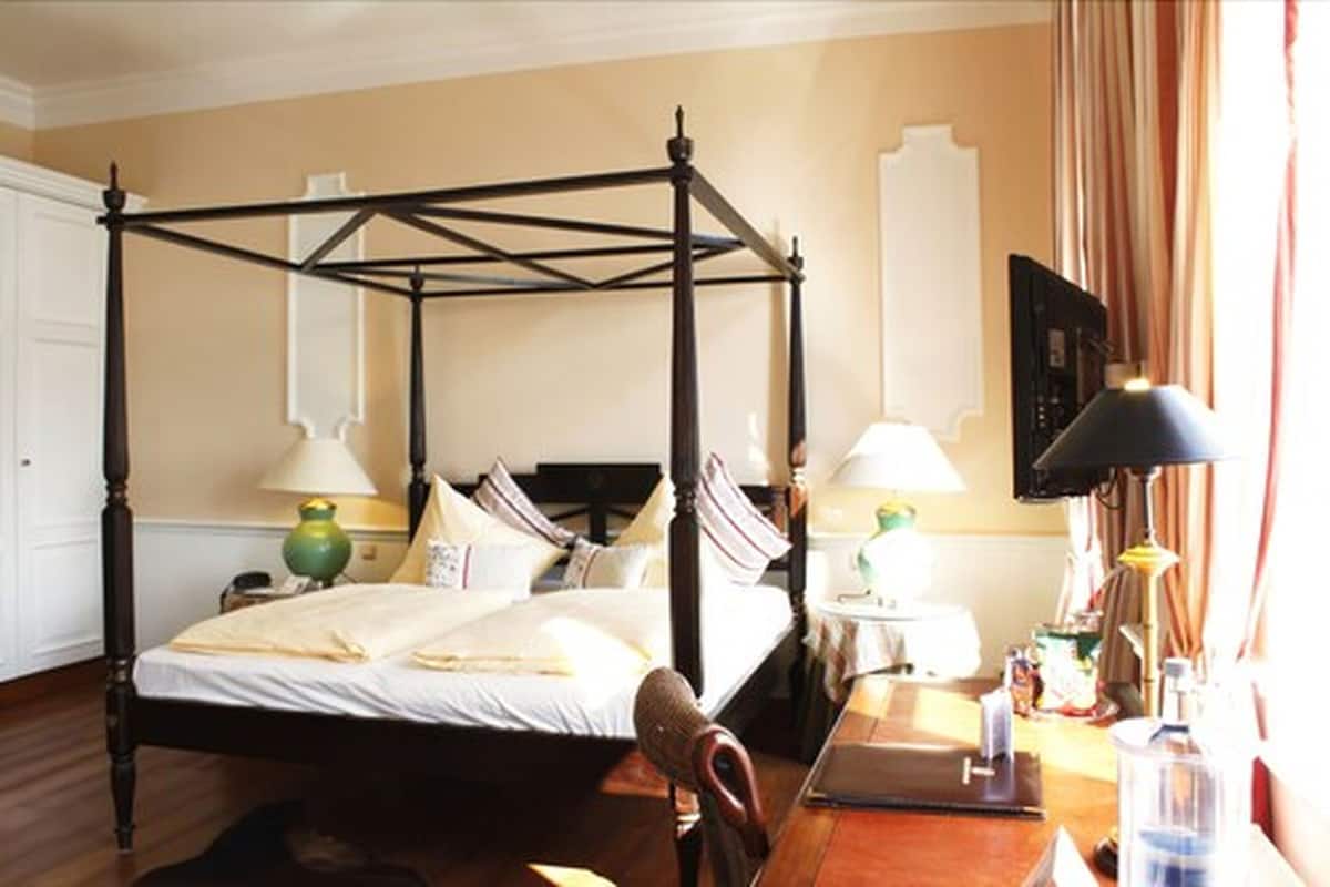Un lit à baldaquin avec un cadre en bois, une table de chevet avec une lampe et un bureau avec un ordinateur et une bouteille.