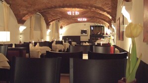A restaurant with a rustic interior, featuring exposed brick ceilings and multiple chandeliers. Tables are set with white tablecloths and candles, and there is a vase with a single yellow tulip on the table. - Hotel Lehmeier (Neumarkt in der Oberpfalz)