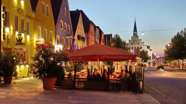 A traditional European street with colorful buildings, a central outdoor seating area, and a church spire in the background. - Hotel Lehmeier (Neumarkt in der Oberpfalz)