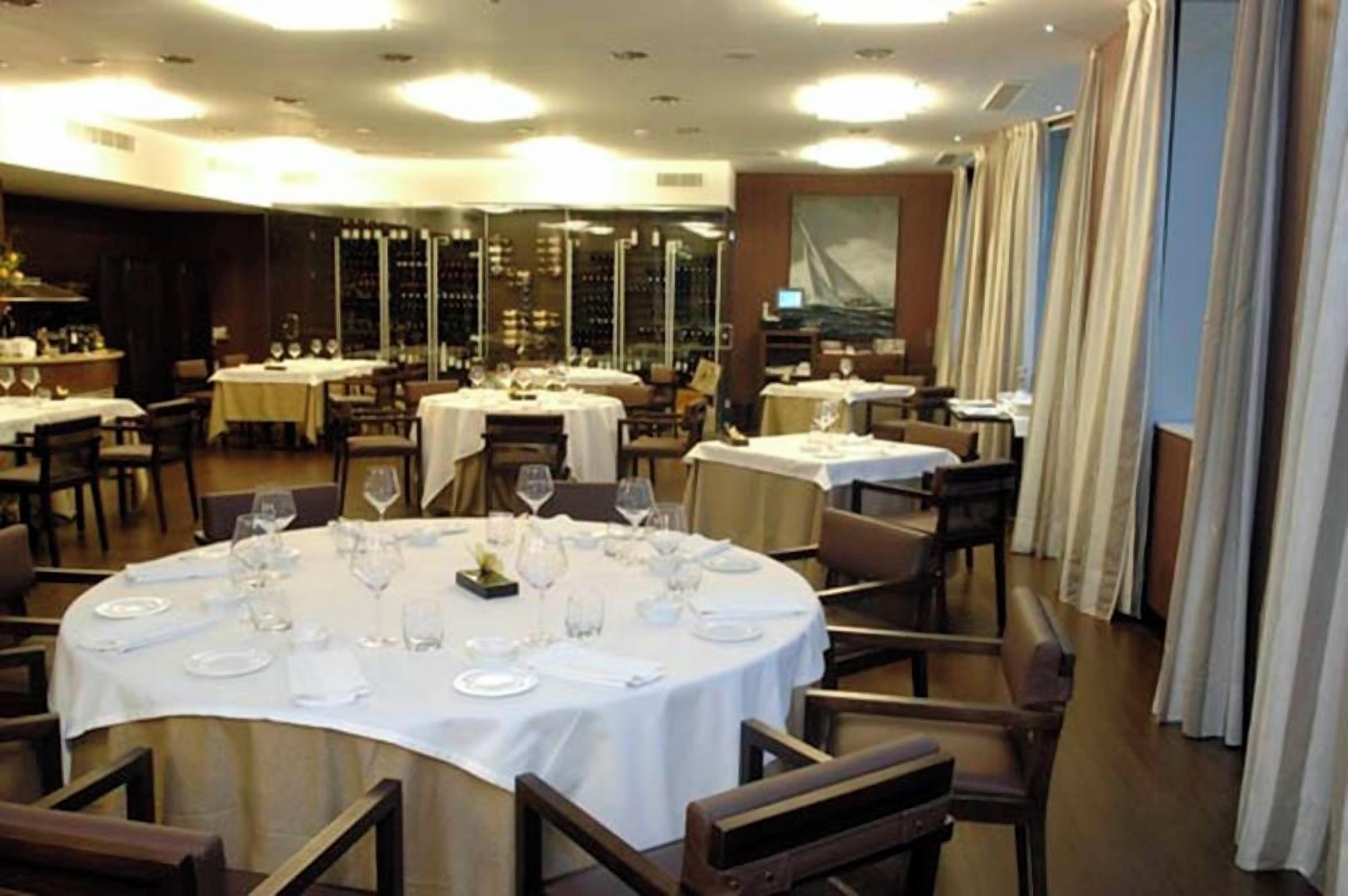 A restaurant with round tables set for dining, a bar area with bottles, and a large painting on the wall.