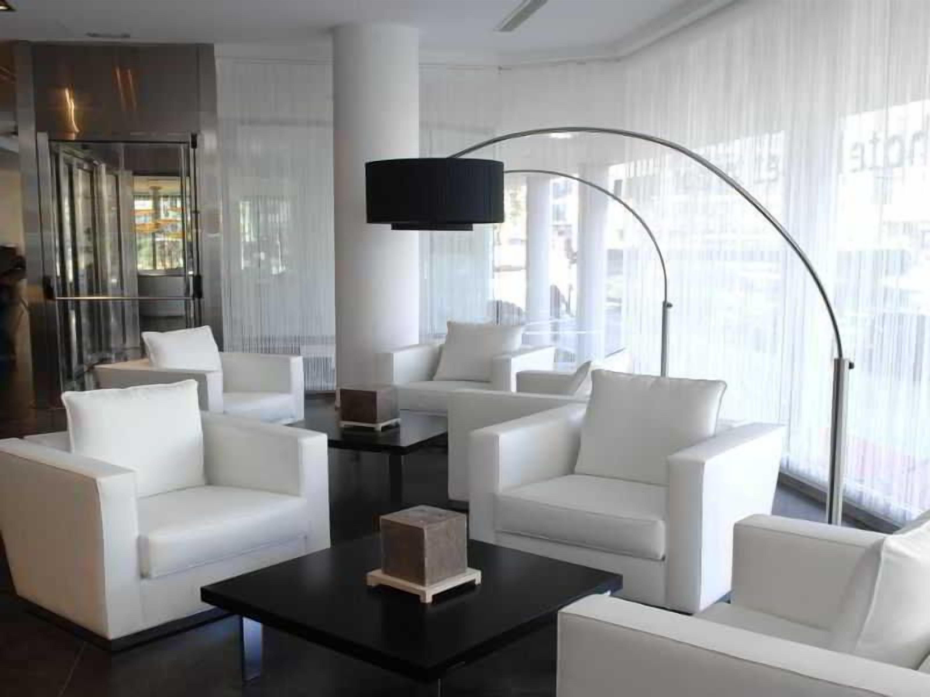 Un moderno vestíbulo de hotel con sofás blancos dispuestos alrededor de una mesa de centro, una lámpara de pie grande y vistas de la ciudad a través de amplias ventanas.
