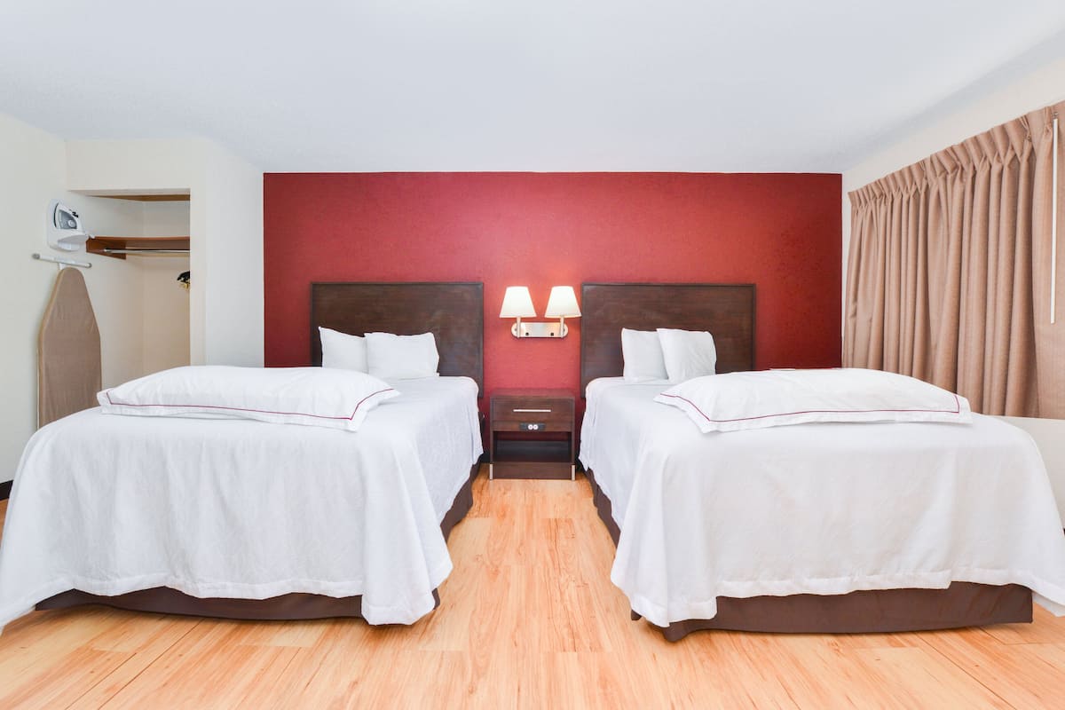 A hotel room with two beds, a wooden floor, a red accent wall, and a nightstand with lamps.
