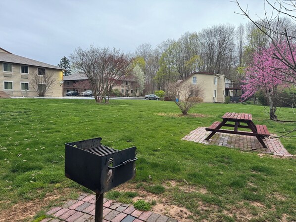 A grassy outdoor area with a barbecue grill, picnic table, and a small tree with pink blossoms. - Red Roof Inn PLUS+ & Suites Guilford (Guilford)