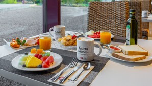 Un desayuno con una mesa para dos, con platos de fruta, tostadas y bebidas, y vista a un espacio verde al exterior.