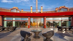 Un edificio moderno con fachada roja, grandes ventanales de vidrio y una zona de estar exterior con mobiliario de mimbre.
