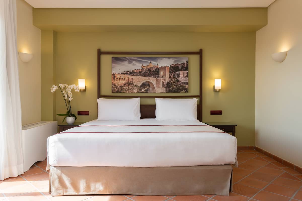 A hotel room with a large bed, two bedside tables, a wall-mounted picture of a historic bridge, and a potted plant.