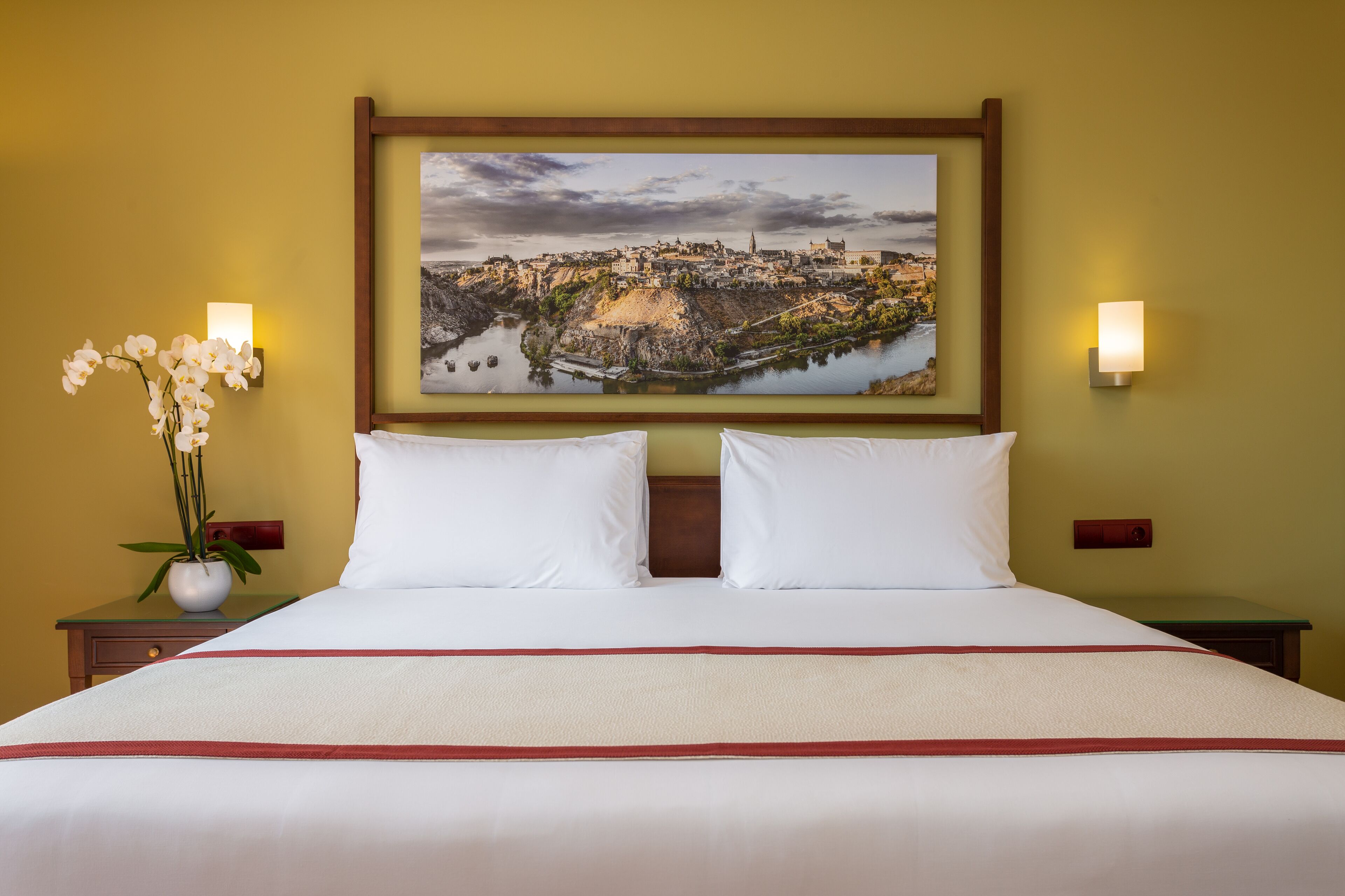 Una cama bien hecha con sábanas blancas y un detalle rojo, un cuadro de paisaje enmarcado encima, y dos lámparas fijadas a la pared a cada lado.