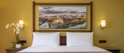 A neatly made bed with white linens and a red accent, a framed landscape painting above it, and two wall-mounted lamps on either side.