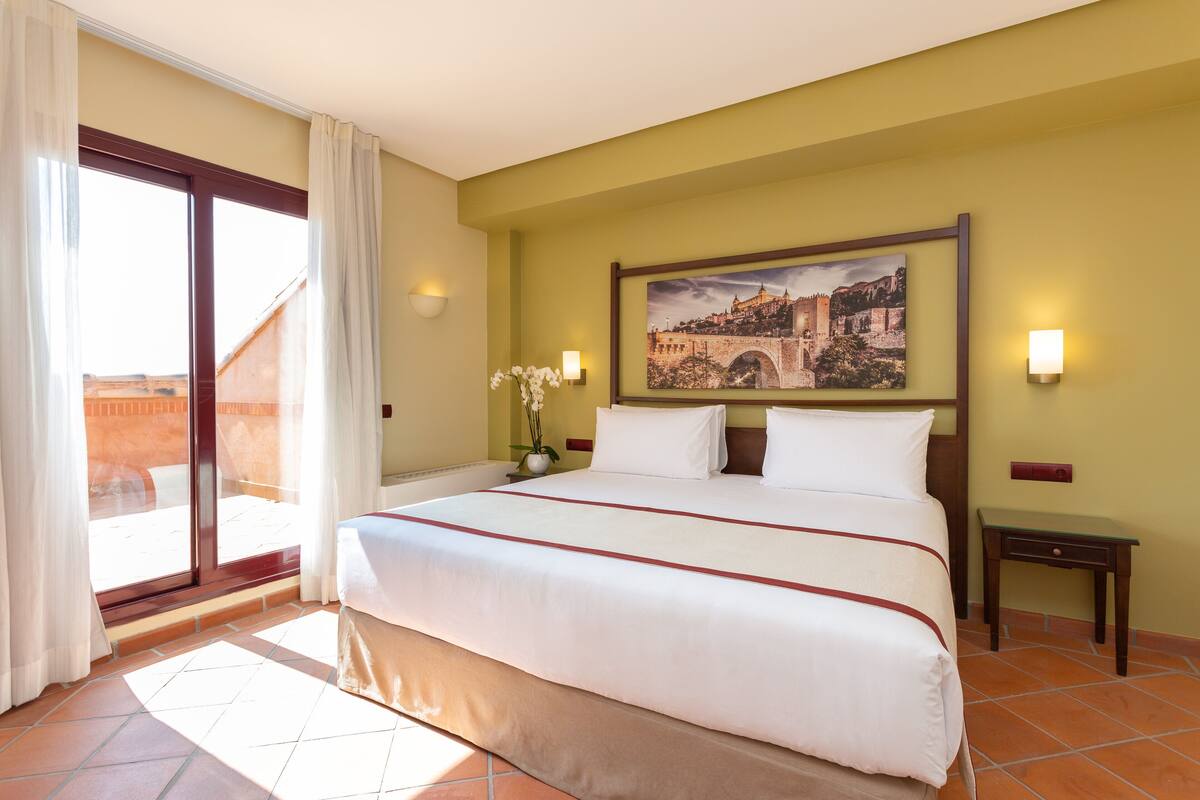A hotel room with a large bed, a view of a cityscape through a sliding glass door, a small table, and a wall-mounted light.