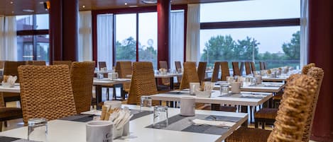 Un restaurant où les tables sont dressées pour le service, avec des chaises en osier et une vue sur l’extérieur.