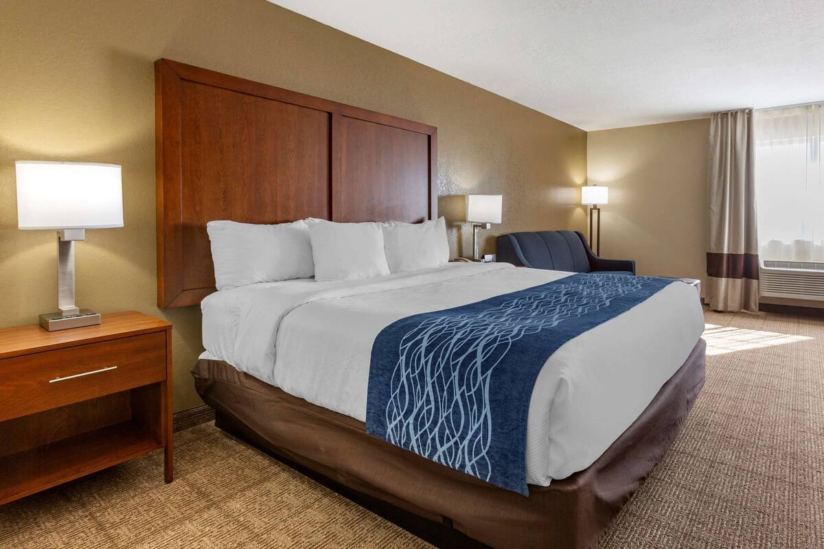 A hotel room with a large bed, wooden headboard, two bedside lamps, a nightstand, and a view of the city through the window.